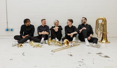 a group of 5 people casually seated on the floor, wearing black outfits, smiling and chatting with brass instruments laid out in front of them