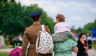 A soldier in uniform walks next to a woman in a vintage dress, with a young child on her shoulders