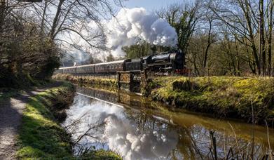 A chugging steam train passes by a river on a sunny day