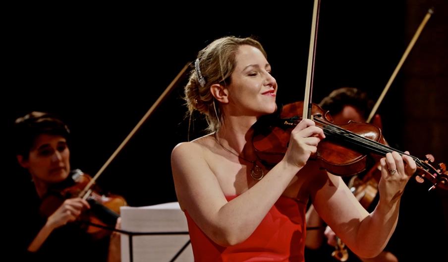 a woman wearing a red gown, holding a violin to play and plucking it with one finger, other violin bows visible in the background