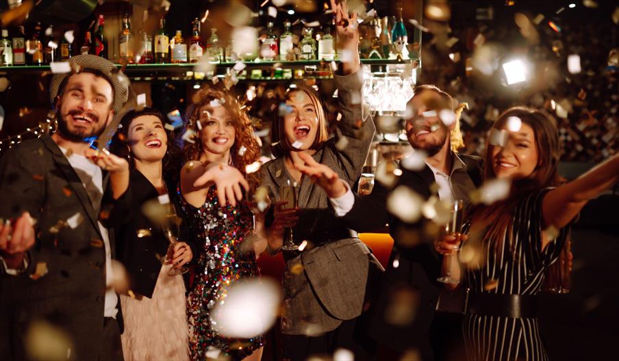 Group of people celebrating at a stylish bar with confetti falling, dressed in formal and festive attire, holding drinks with a backdrop of shelves st