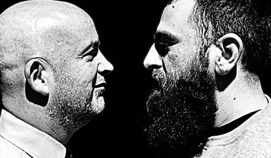 A black and white image of two men staring at each other in a face off. The man on the right has a long bushy beard. The man on the left is bald.