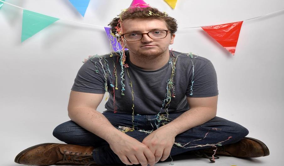 Image of a man sitting on the floor, covered in streamers and wearing a party hat