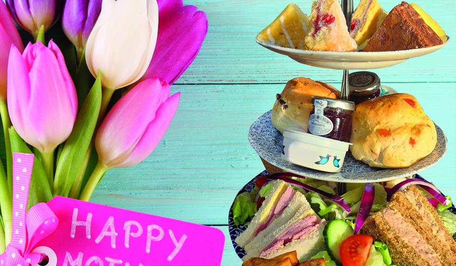 An image of a Mother's Day Afternoon Tea, against a light blue background, next to a bunch of tulips with Happy Mother's Day written on the card