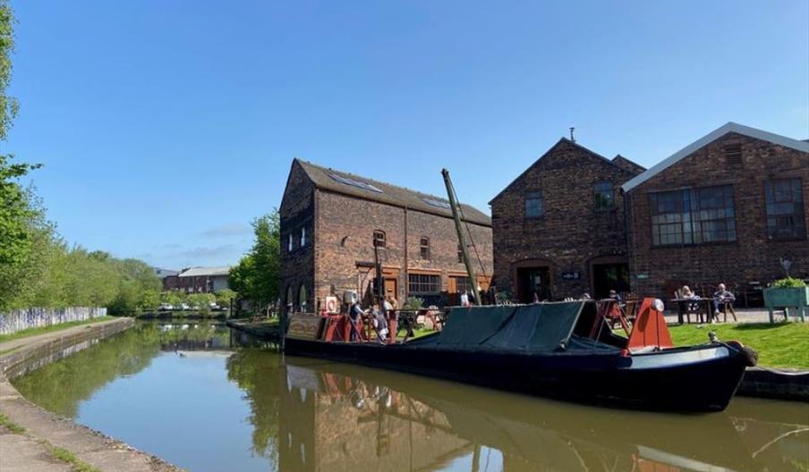 An historic narrowboat, moored up beside some canalside heritage buildings, on a sunny day