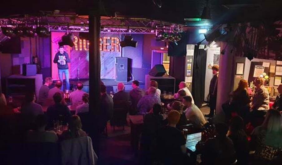 Image shows a comedian on stage at The Rigger, in front of a packed audience