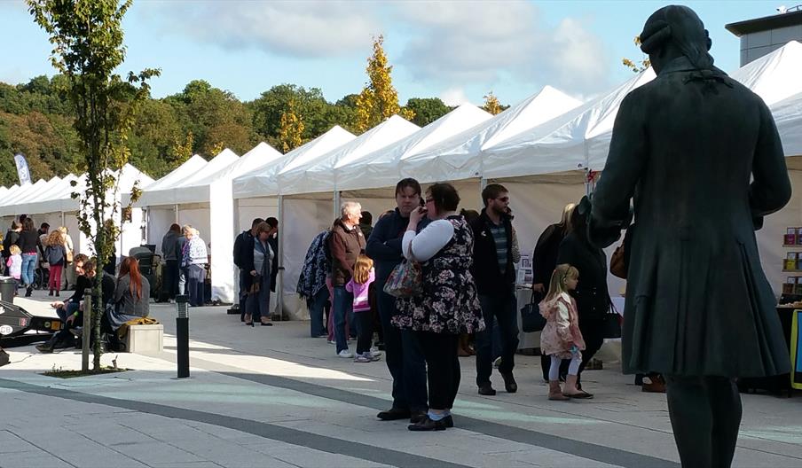 Image shows a row of white gazebos with market stalls inside them, with shoppers milling around in the sunshine, and a statue of Josiah Wedgwood takin