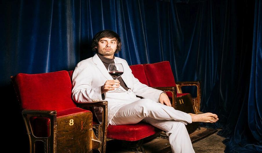 Image shows a man sitting in an old theatre/cinema chair, with a glass of red wine
