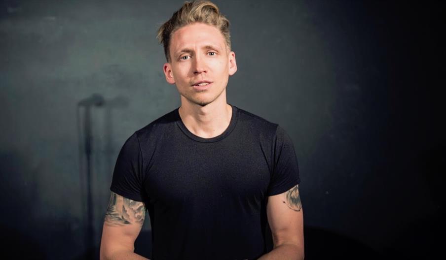 A man in a black t-shirt looks at the camera. It looks like he is performing on a stage but you can't see the audience, only a microphone shadow