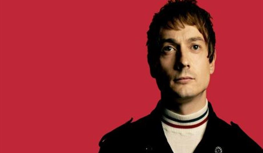 Image shows the top half of a man - one of the comedians on the line-up - standing against a red backdrop