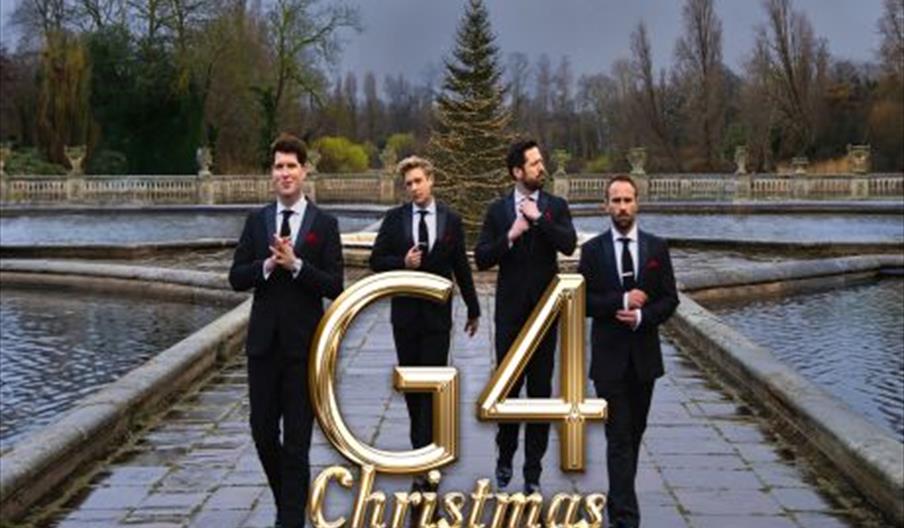 Four men in black suits and ties walk beside a lake at Christmastime. There is a Christmas tree behind them, and G4 Christmas layered on top of the im