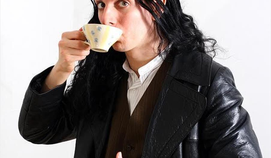 An image of a man, with long black hair and a black leather jacket, drinking a cup of tea