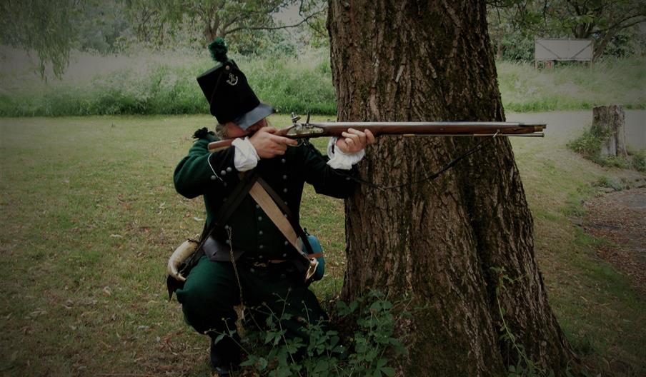 A costumed performer, as a rifleman, crouches behind a tree and takes aim