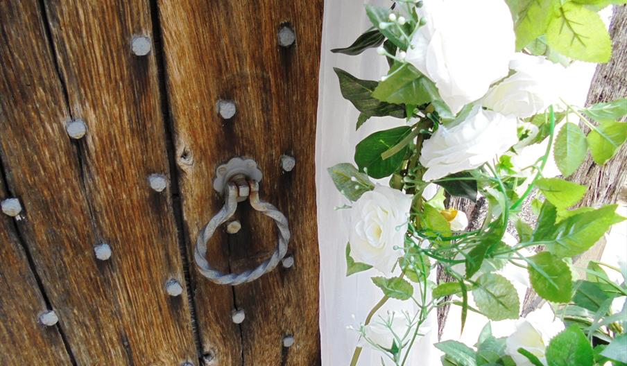 Image shows a close-up of a very old wooden door, with some white roses growing next to it