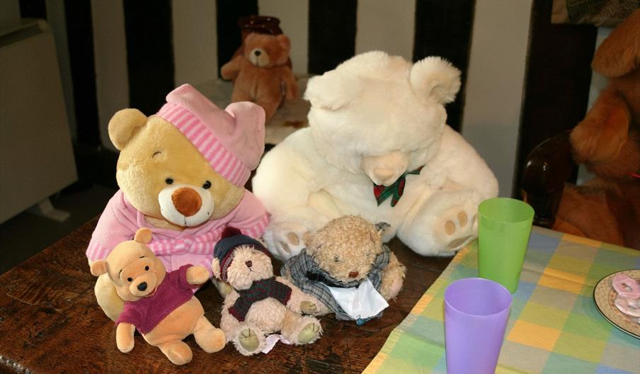 A selection of teddies of different sizes, on a wooden table, with a picnic off to the side