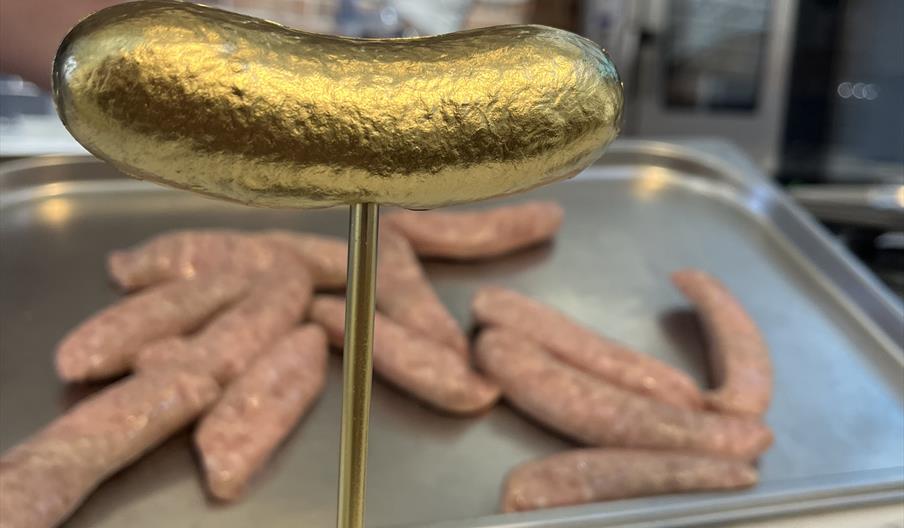 Award trophy shaped like a sausage in gold with trays of fresh sausages in the background.