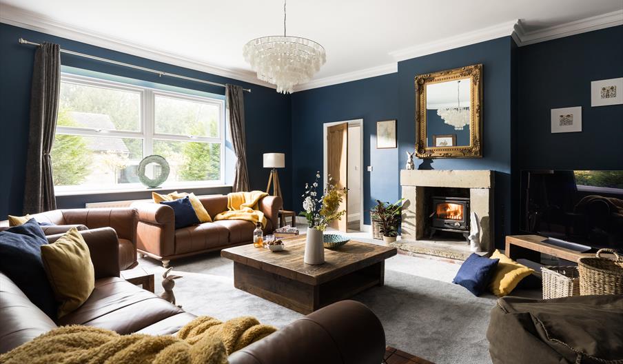 A spacious lounge area, with large sofas, a roaring log fire, and a large window looking into the garden
