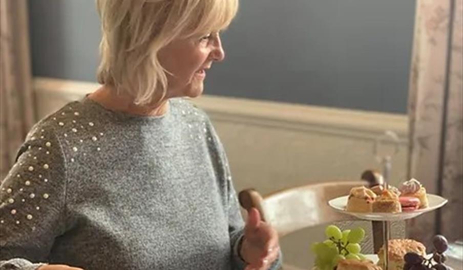 A lady prepares to tuck into afternoon tea, gazing at the cakes on the stand, with her hand on a teapot