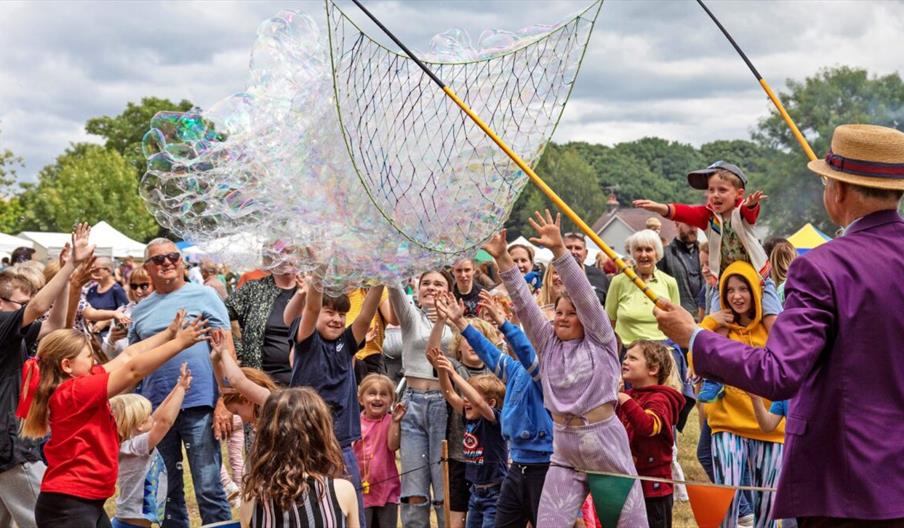 A man with a huge net creates hundreds of bubbles in front of a crowd of people, mostly children