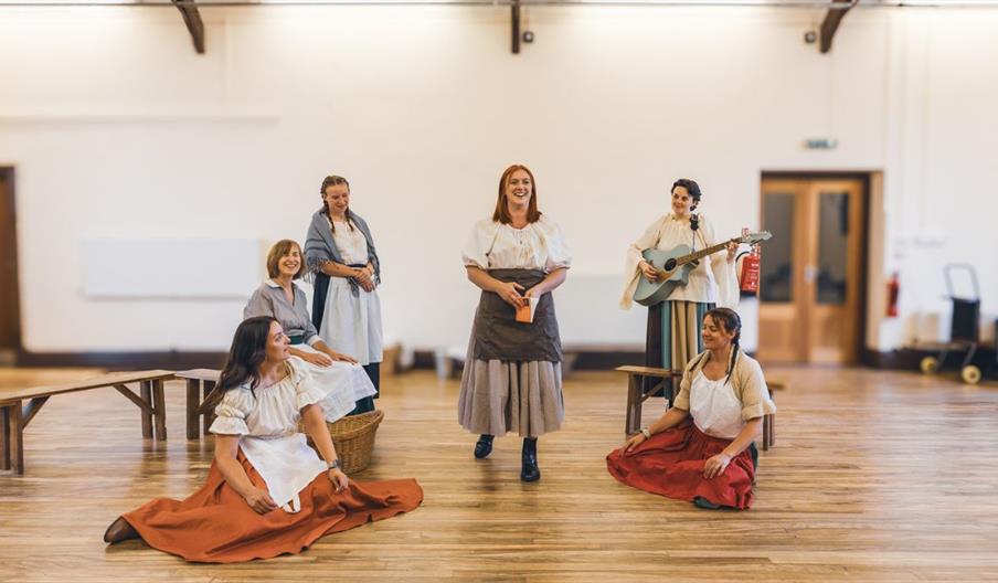 Six ladies inside a room with a wooden floor, one holding a guitar and two clapping