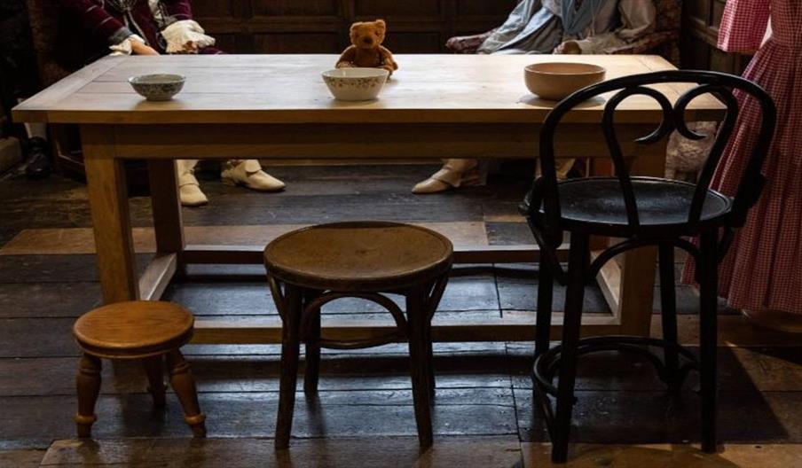 A wooden table set with bowls and a small teddy bear, surrounded by mismatched stools and chairs on an old wooden floor inside a historic room decorat
