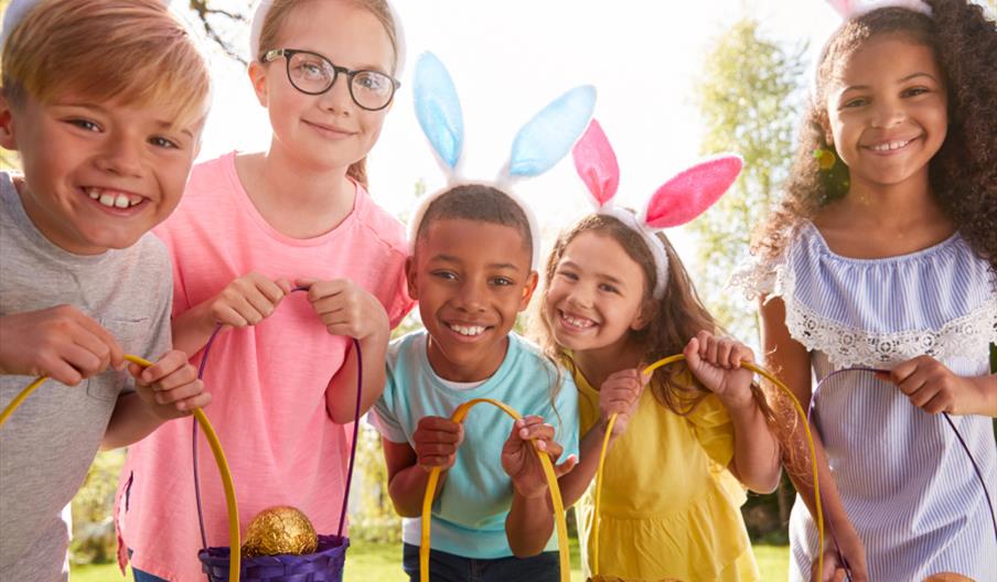 A group of children with Easter eggs in baskets, wearing bunny ears, smile at the camera