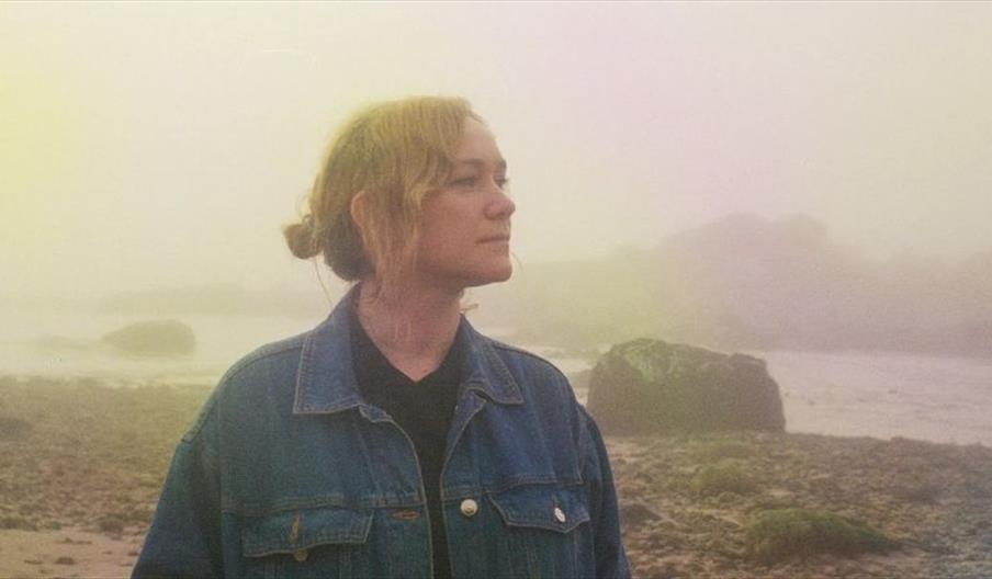 Image shows a woman, standing by a river on a foggy day, gazing into the distance