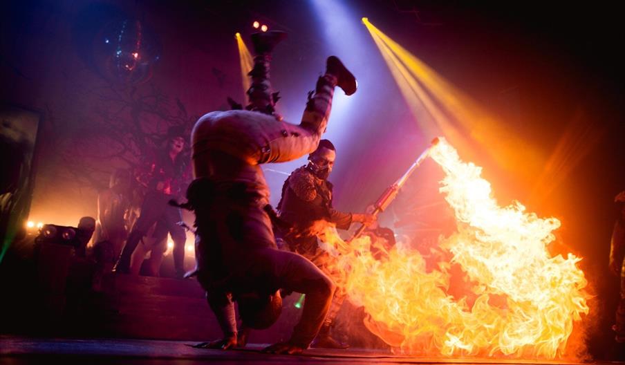Image shows circus performers on stage, with one doing a somersault and another with a flame-thrower