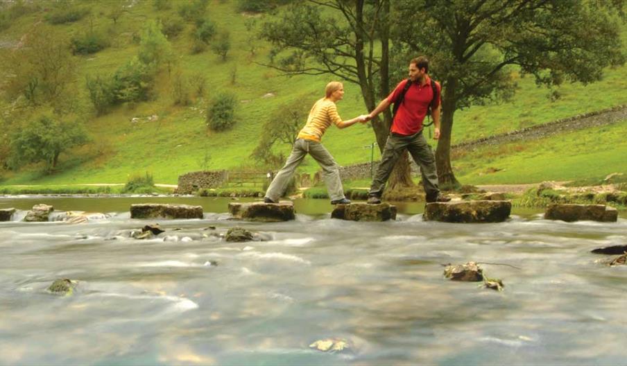 Couple crossing the famous Stepping Stones across the River Dove, Dovedale, Peak District. Staffordshire / Derbyshire border. Image courtesy Tony Plea