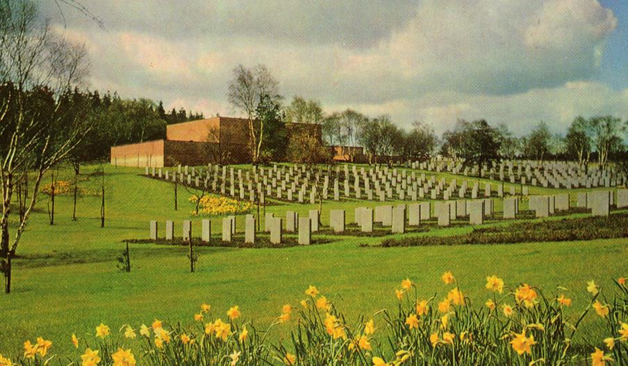 Image shows a cemetery on a sunny day, with rows of graves, daffodils in the foreground, and a building in the background
