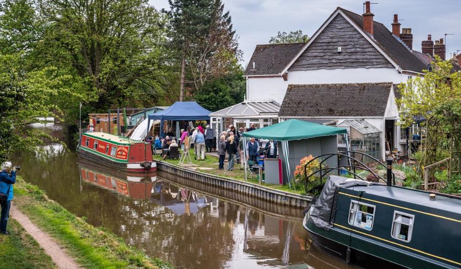 A  canalside garden event with people gathered under gazebos beside two moored narrowboats. One boat is painted red and green with the name "The Admir