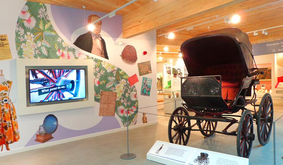 A well-lit area with a large carriage and a plasma screen on a colourfully-decorated wall featuring wallpaper patterns, gramophone, costumes and famou