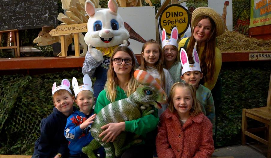 Image shows a group of visitors, some wearing bunny ears, and an Easter Bunny in the background, while a member of staff cradles a baby 'dinosaur'