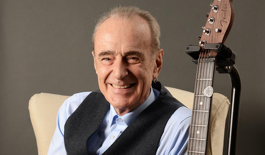 Francis Rossi smiles, sitting next to a guitar