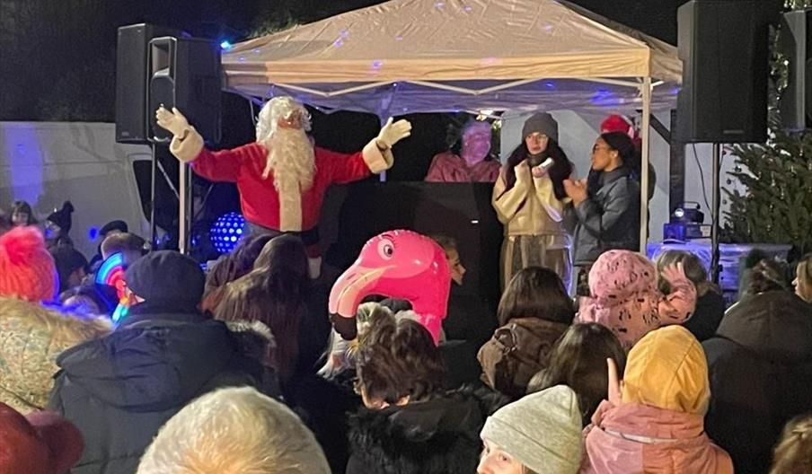 A crowd of people watch on as Santa speaks, standing under a gazebo with some singers and a pianist or DJ