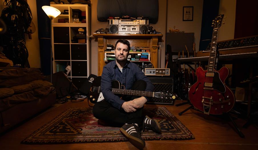 Image shows Jim Moray sitting down in his lounge with his guitars and other musical paraphernalia