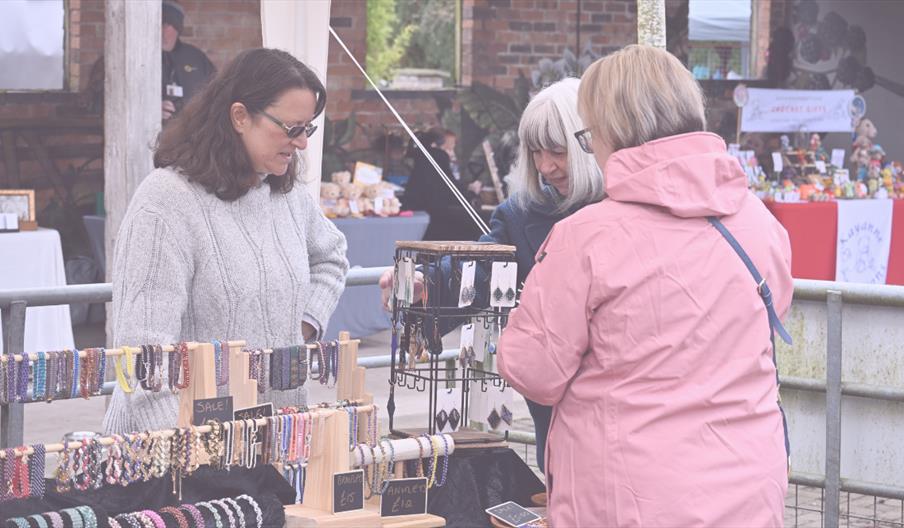 An outdoor craft market stall displaying rows of handmade bracelets and earrings on wooden and metal stands, while three people stand close together b