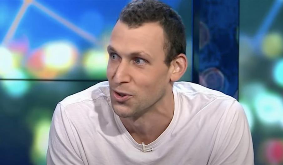 Michael Shafar wearing a plain white T-shirt seated indoors against a colorful blurred background with circular light patterns.