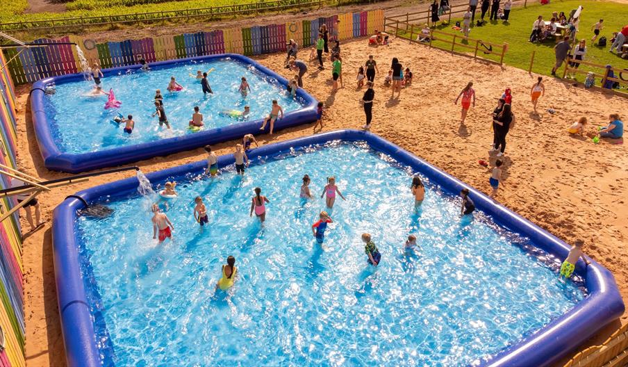 An overhead shot of children playing in two huge paddling pools, and others on a beach