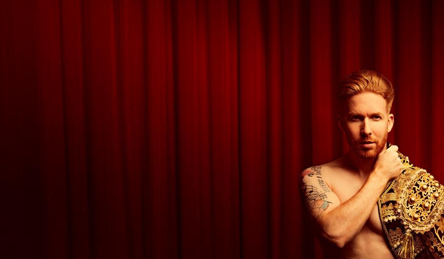 A topless man, with a Spanish matador's jacket draped over his shoulder, looks at the camera as he stands in front of a red curtain