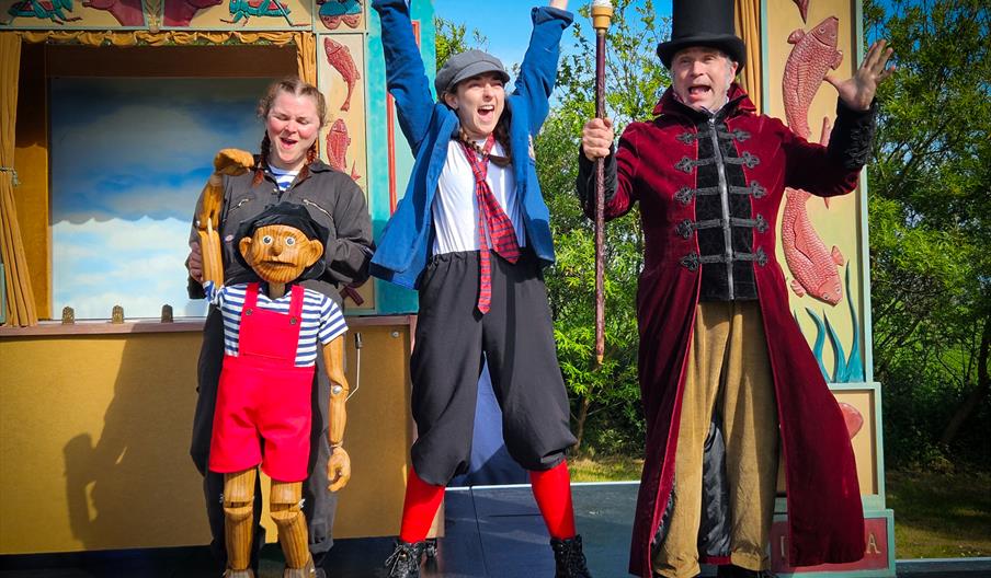 three people on stage in costume dress. One person is holding a wooden puppet called Pinocchio