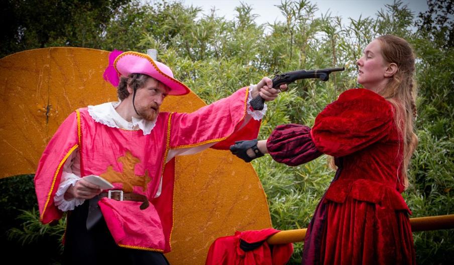 image A: two people in costume dress acting out a fighting seen from the musketeers using pretend pistols 
Image B: two people in costume dress acting