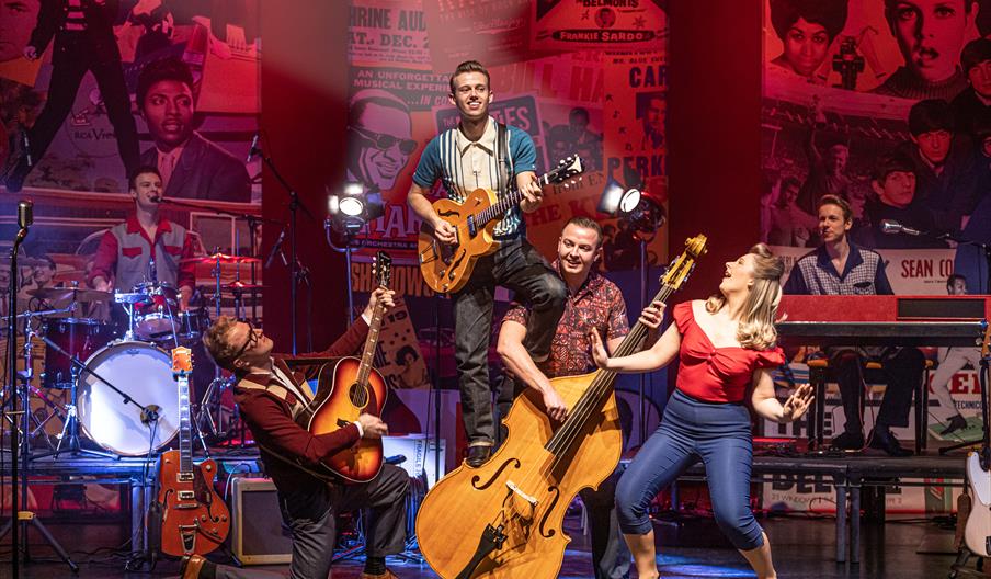 A band plays music on stage, against a backdrop of images and newspaper cuttings from the 50s and 60s