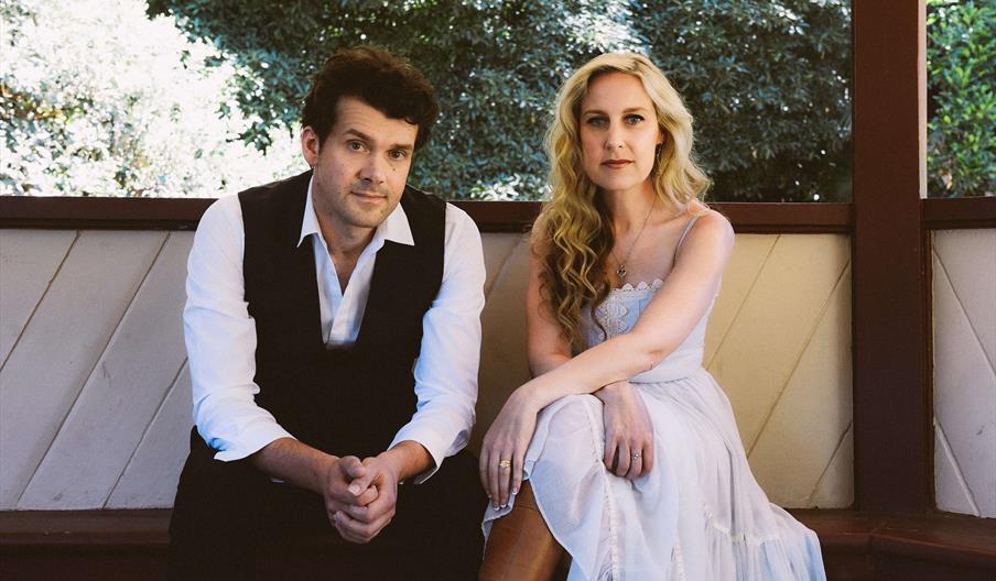 A man dressed in black and white and a woman in a long white dress, sitting in a wooden bandstand or similar