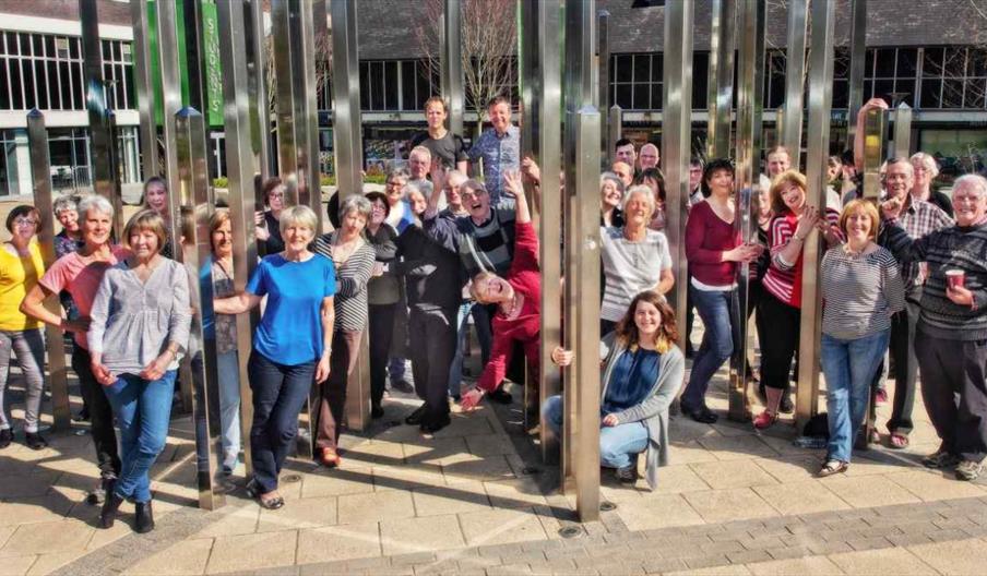 Large group of people gathered outdoors among tall metal columns, posing together in a public square near modern buildings.