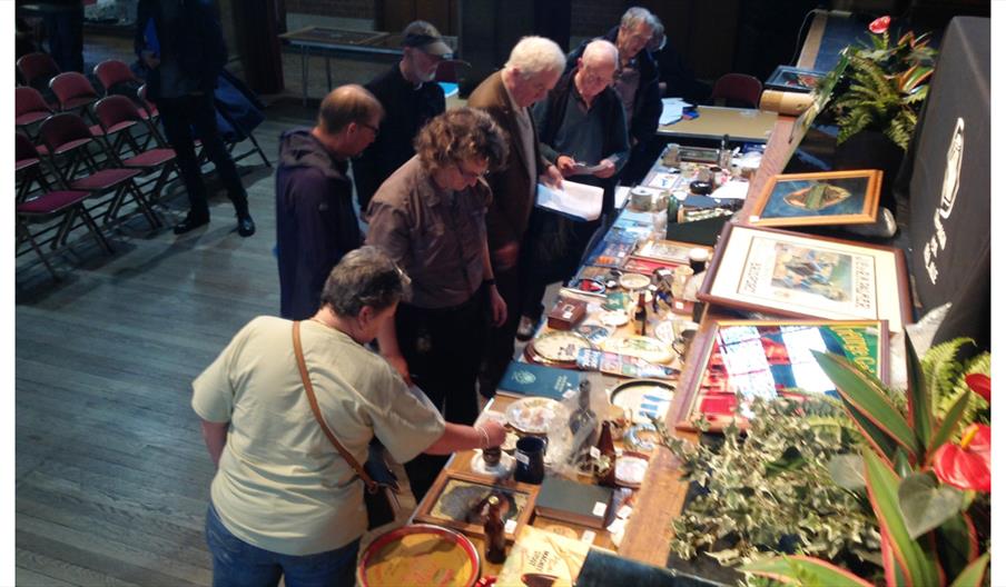 People browsing tables of vintage brewery memorabilia and collectibles at an indoor auction event.