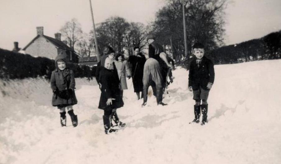 Historic black-and-white photograph showing children and adults walking through deep snow on a rural road near houses and trees.