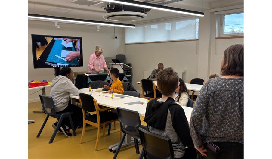 Participants take part in a bookbinding workshop while an instructor demonstrates techniques at the front of the room.