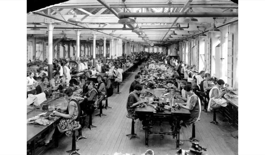 Historic factory workshop with rows of workers seated at long tables.