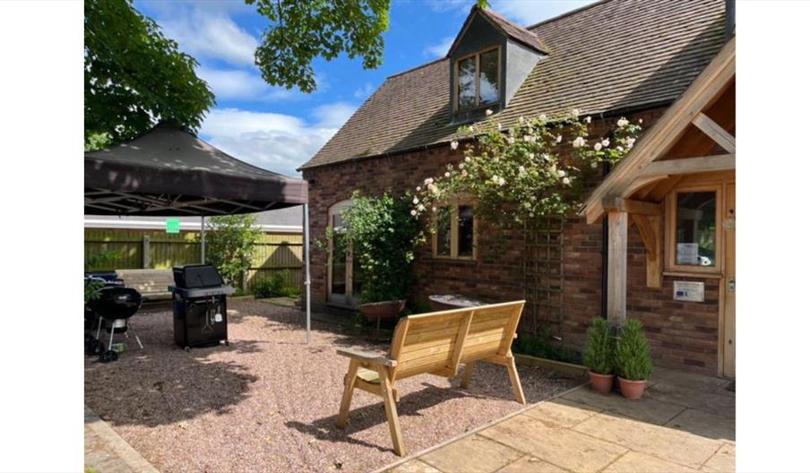 Brick cottage courtyard with outdoor seating, BBQ grills, and a canopy in a sunny garden setting.